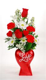 Red ceramic heart vase filled with two long stem red roses, white spray daisies, valentine red carnations, baby breath, and mixed greenery and two red valentine hearts.